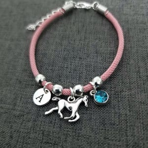 K&ouml;nnte beinhalten: Rosa geflochtenes Armband mit silbernen Perlen, einem silbernen Pferdeanh&auml;nger, einem silbernen Anh&auml;nger mit dem Buchstaben A und einem hellblauen Kristallanh&auml;nger.