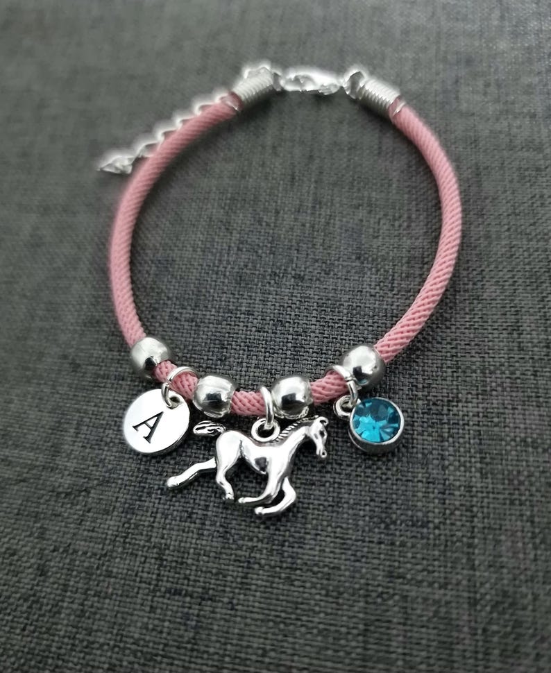 K&ouml;nnte beinhalten: Rosa geflochtenes Armband mit silbernen Perlen, einem silbernen Pferdeanh&auml;nger, einem silbernen Anh&auml;nger mit dem Buchstaben A und einem hellblauen Kristallanh&auml;nger.
