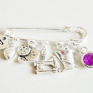 May include: Silver charm brooch with a sewing theme. The brooch features a sewing machine, spool of thread, buttons, a heart, and a purple rhinestone. The brooch also has a charm with the letter "E" on it.