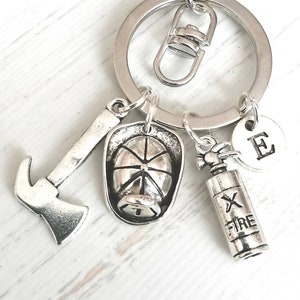 May include: Silver keychain with a firefighter theme. It features a miniature axe, a fire helmet, a fire extinguisher, and a charm with the letter "E".