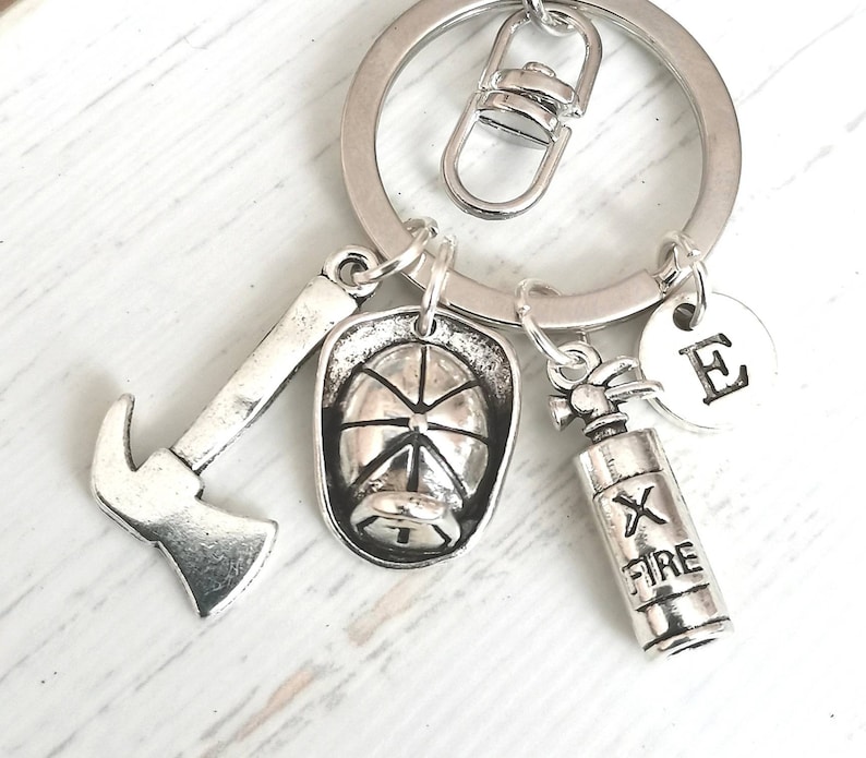 May include: Silver keychain with a firefighter theme. It features a miniature axe, a fire helmet, a fire extinguisher, and a charm with the letter "E".