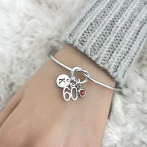 K&ouml;nnte beinhalten: Ein silbernes Armreif-Armband mit einem Knotendesign und drei Anh&auml;ngern. Die Anh&auml;nger sind ein runder Anh&auml;nger mit dem Buchstaben "K" darauf, ein Anh&auml;nger mit der Zahl "60" darauf und ein kleiner Anh&auml;nger aus rotem Edelstein.