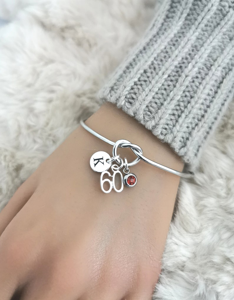K&ouml;nnte beinhalten: Ein silbernes Armreif-Armband mit einem Knotendesign und drei Anh&auml;ngern. Die Anh&auml;nger sind ein runder Anh&auml;nger mit dem Buchstaben "K" darauf, ein Anh&auml;nger mit der Zahl "60" darauf und ein kleiner Anh&auml;nger aus rotem Edelstein.