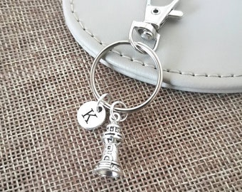 May include: Silver keychain with a lighthouse charm and a small round charm with the letter K. The keychain is on a brown woven surface.