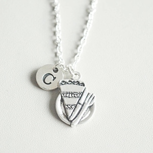 Puede incluir: Un collar de plata con un dije de un plato con una rebanada de pastel y un tenedor. El collar también tiene un dije de plata con la letra C.