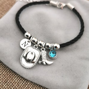May include: A black braided bracelet with silver charms, including a cowboy hat, a boot, a turquoise blue gemstone, and a charm with the letter N.