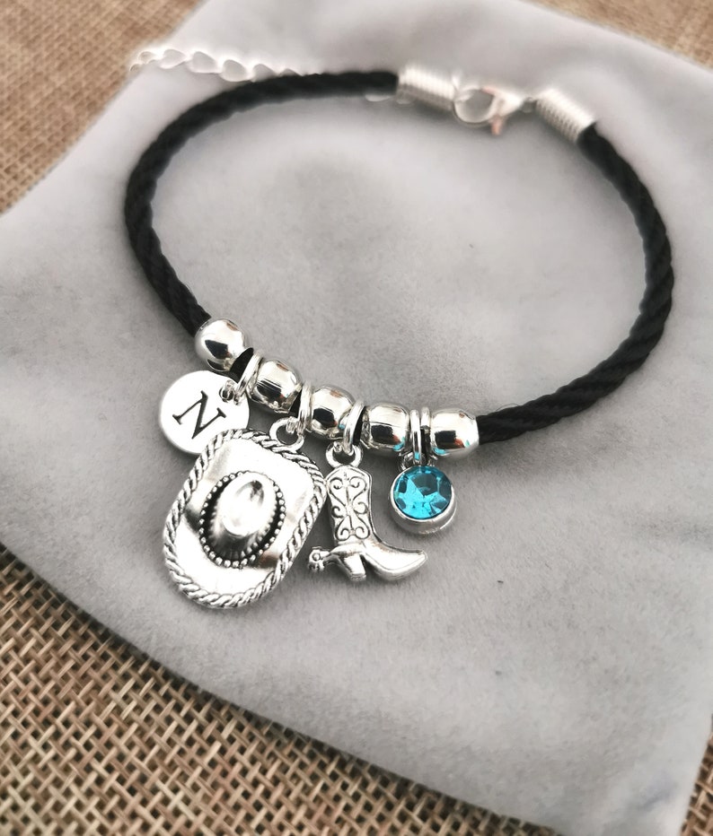 May include: A black braided bracelet with silver charms, including a cowboy hat, a boot, a turquoise blue gemstone, and a charm with the letter N.