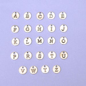 Op de afbeelding: Een set van 26 zilverkleurige metalen alfabetletterbedels. Elke bedel is een cirkel met een verhoogde letter in het midden. De letters zijn alfabetisch gerangschikt.