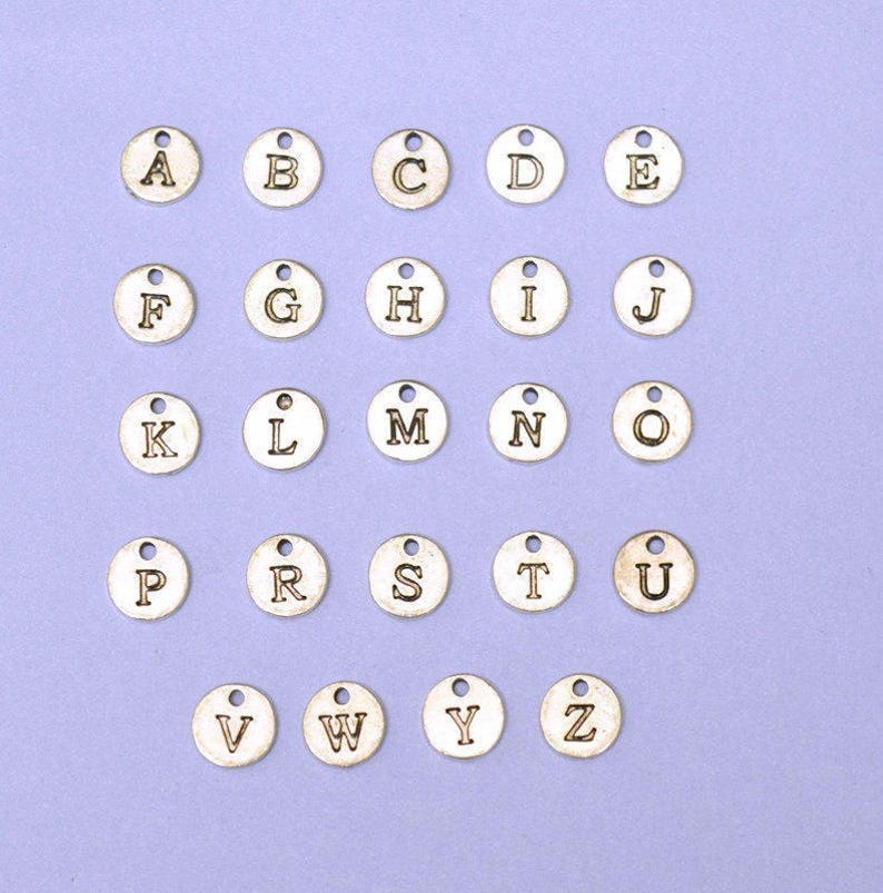 Op de afbeelding: Een set van 26 zilverkleurige metalen alfabetletterbedels. Elke bedel is een cirkel met een verhoogde letter in het midden. De letters zijn alfabetisch gerangschikt.