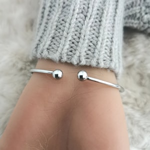 May include: A silver-coloured, open cuff bracelet with a small ball at each end. The bracelet is worn on a wrist, with a grey sweater in the background. The bracelet is a simple, elegant piece of jewellery.