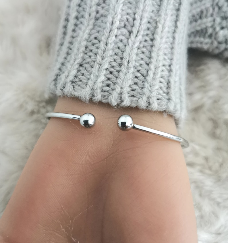 May include: A silver-coloured, open cuff bracelet with a small ball at each end. The bracelet is worn on a wrist, with a grey sweater in the background. The bracelet is a simple, elegant piece of jewellery.