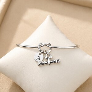 May include: A silver-colored bangle bracelet with a knot design. The bracelet has three charms: a round charm with the letter "A", a graduation cap charm, and a charm that reads "GRADUATE".