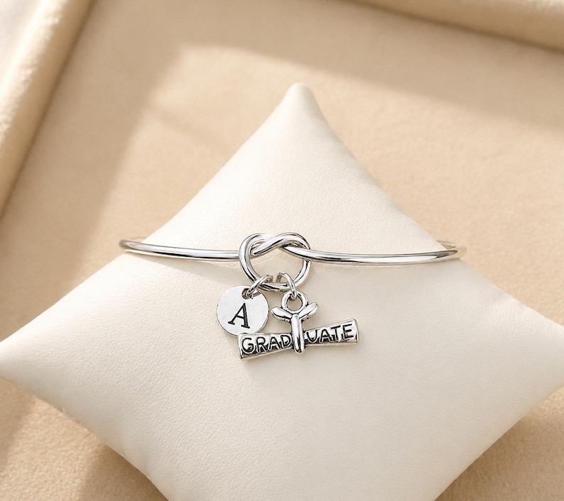 May include: A silver-colored bangle bracelet with a knot design. The bracelet has three charms: a round charm with the letter "A", a graduation cap charm, and a charm that reads "GRADUATE".