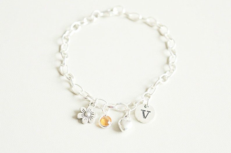May include: A silver charm bracelet with a flower, a heart, a small gemstone, and a charm with the letter "V" hanging from it.