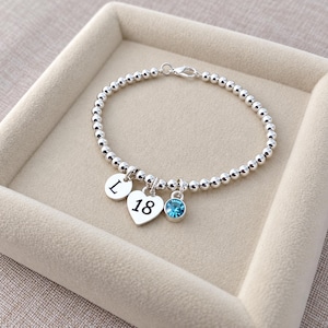 May include: A silver-toned beaded bracelet featuring three dangling charms: a circular disc engraved with the letter L, a heart-shaped charm engraved with the number 18, and a small circular charm set with a single bright blue crystal gemstone.