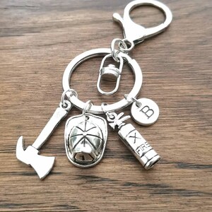 May include: Silver keychain with a firefighter theme. It features a fire hat, axe, fire extinguisher, and a charm with the letter "B".
