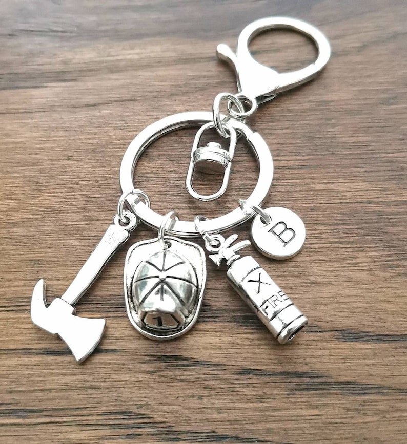 May include: Silver keychain with a firefighter theme. It features a fire hat, axe, fire extinguisher, and a charm with the letter "B".