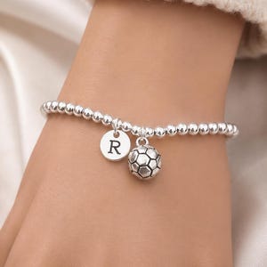 May include: Silver beaded bracelet with a soccer ball charm and a round charm with the letter "R". The bracelet is worn on a wrist, and the background is a soft, neutral color.