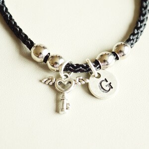 Può includere: Braccialetto in corda intrecciata nera con perline argentate e un ciondolo a forma di chiave con un cuore e ali, e un disco argentato con la lettera 'G' su di esso.