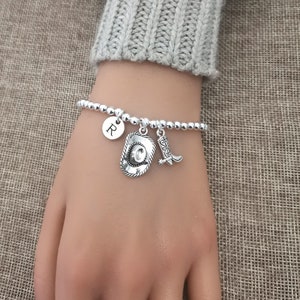 May include: Silver beaded bracelet with cowboy hat, boot, and initial charms. The bracelet features a small round charm with the letter "R". The charms are detailed and add a Western flair.