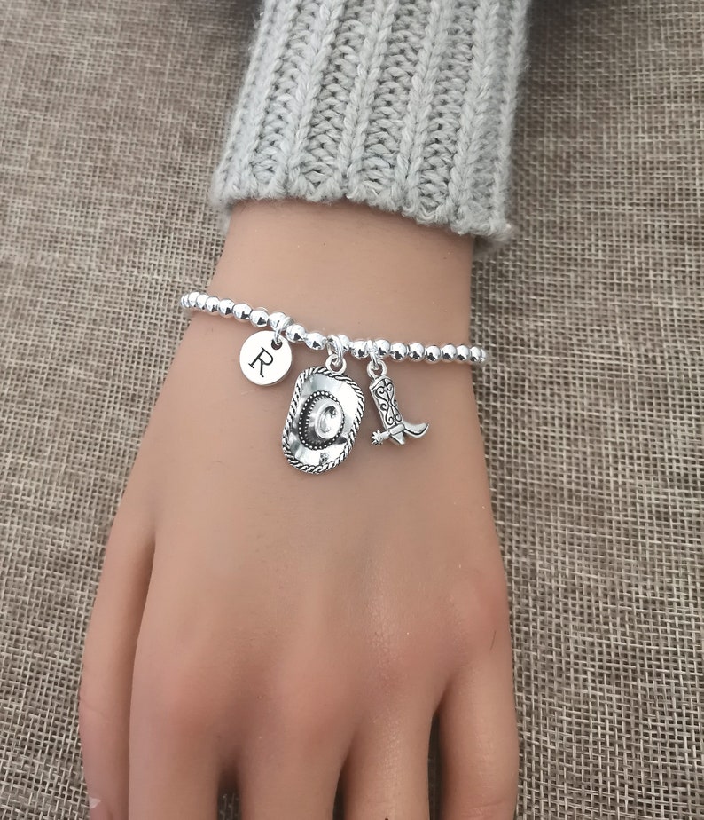 May include: Silver beaded bracelet with cowboy hat, boot, and initial charms. The bracelet features a small round charm with the letter "R". The charms are detailed and add a Western flair.