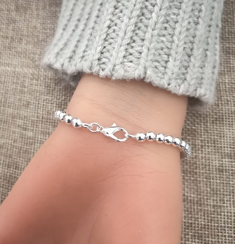 May include: A silver beaded bracelet with a lobster clasp closure. The bracelet is composed of small, round beads. The bracelet is shown on a wrist, with a gray sweater in the background.