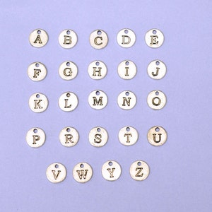 May include: A set of 26 silver-toned metal alphabet letter charms. Each charm is round and features a single letter of the alphabet in a black font. The charms are arranged in alphabetical order on a light purple background.