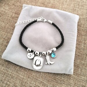 May include: A black cord bracelet with silver charms, including a cowboy hat, a boot, a letter 'N' charm, and a blue rhinestone.