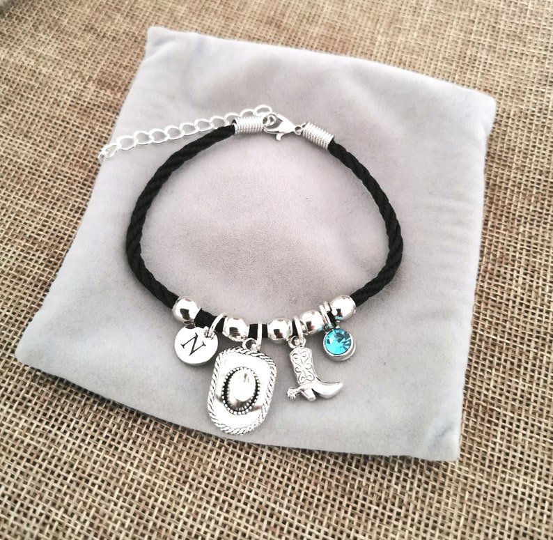 May include: A black cord bracelet with silver charms, including a cowboy hat, a boot, a letter 'N' charm, and a blue rhinestone.