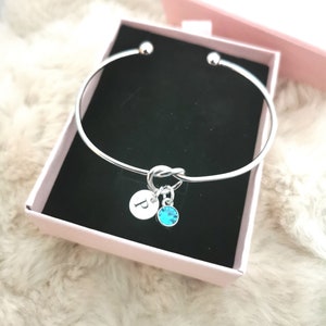 May include: Silver bangle bracelet with a knot charm and a small charm with the letter "P" and a turquoise crystal.