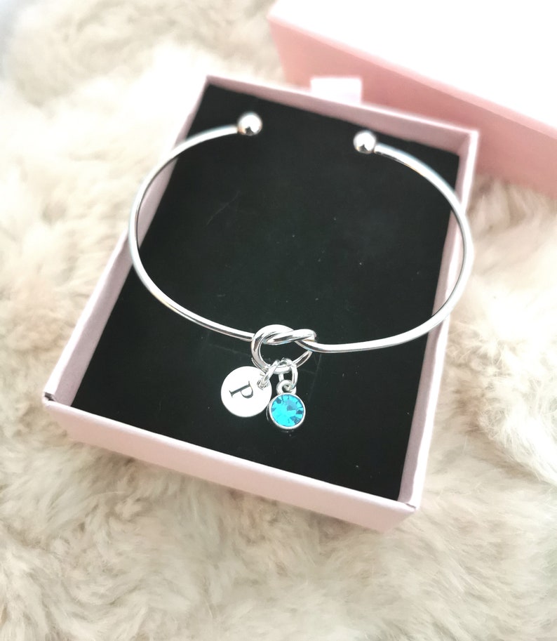 May include: Silver bangle bracelet with a knot charm and a small charm with the letter "P" and a turquoise crystal.