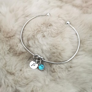 May include: A silver bangle bracelet with a knot detail and two charms. One charm is a round disc with the letter "P" engraved on it. The other charm is a round blue crystal.