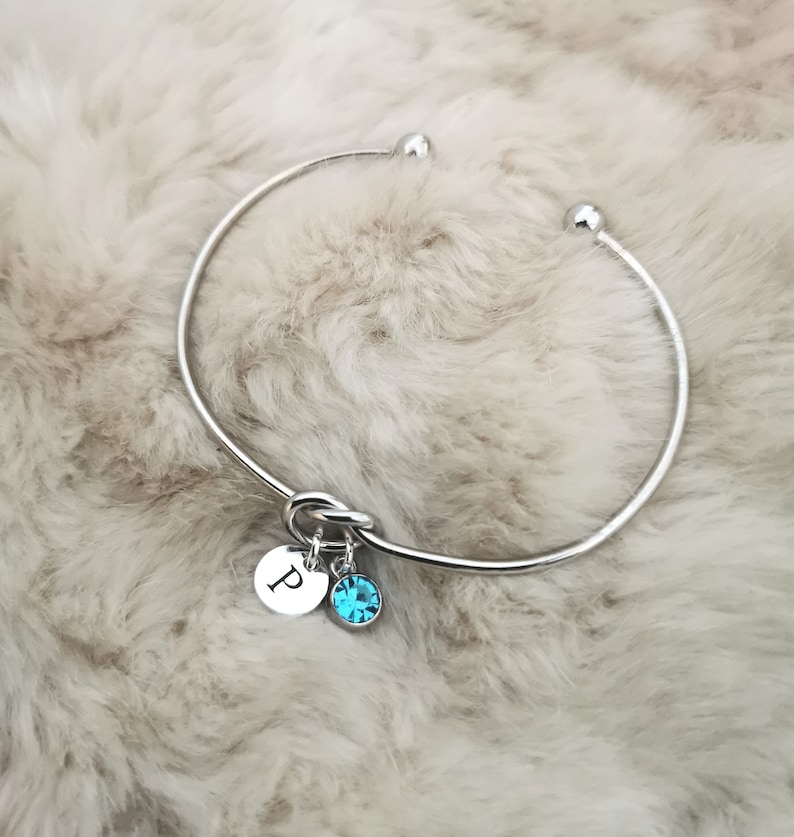 May include: A silver bangle bracelet with a knot detail and two charms. One charm is a round disc with the letter "P" engraved on it. The other charm is a round blue crystal.