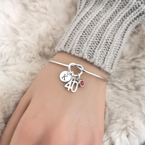 May include: Silver bangle bracelet with a knot design. It features charms including a disc with the letter "K", the number "40", and a small red gemstone. The bracelet is shown on a wrist.