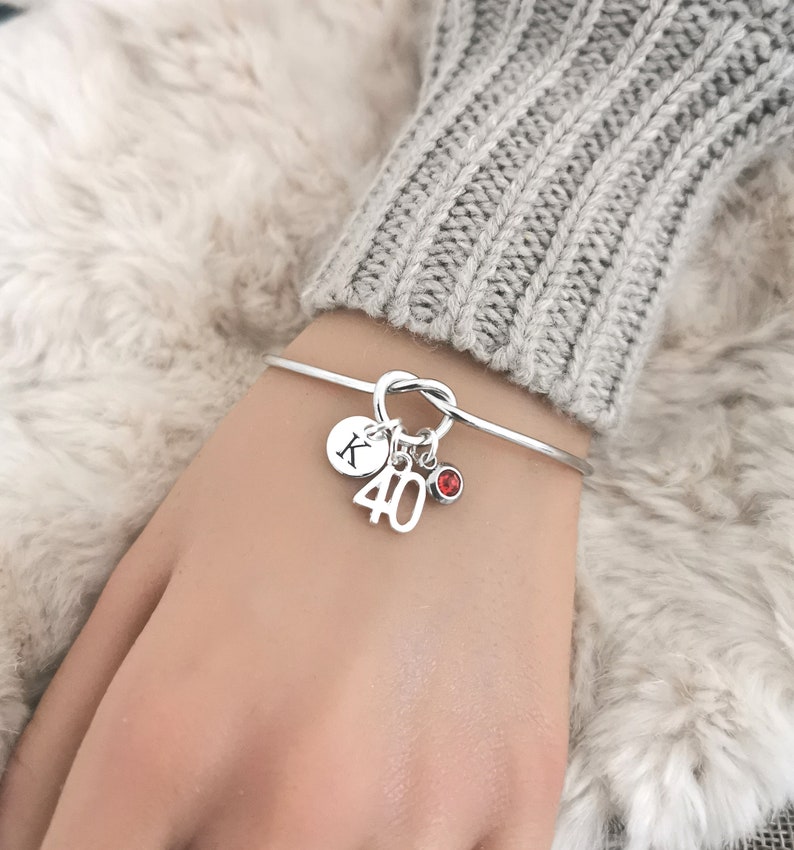May include: Silver bangle bracelet with a knot design. It features charms including a disc with the letter "K", the number "40", and a small red gemstone. The bracelet is shown on a wrist.