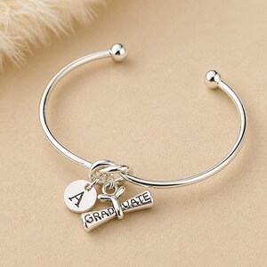 May include: A silver-colored bangle bracelet with a round charm engraved with the letter "A" and a graduation scroll charm that reads "GRADUATE". The bracelet has a knot design and ball ends.