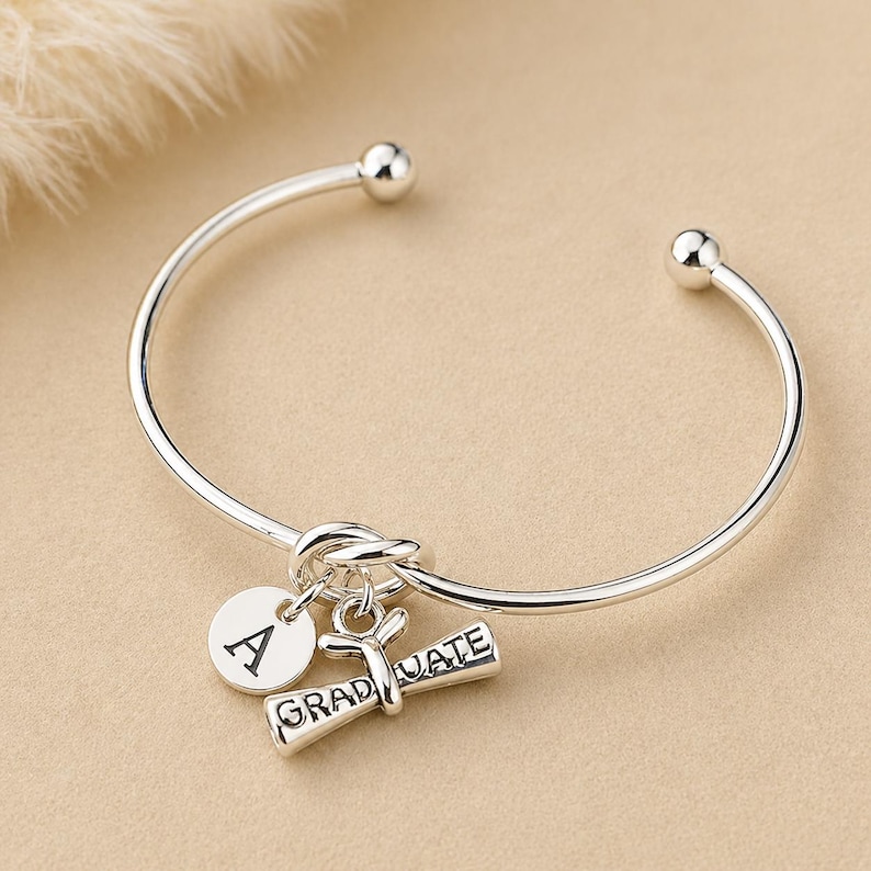 May include: A silver-colored bangle bracelet with a round charm engraved with the letter "A" and a graduation scroll charm that reads "GRADUATE". The bracelet has a knot design and ball ends.