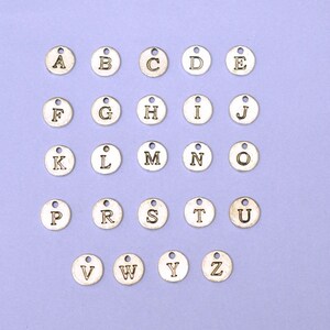 May include: A set of 26 silver-toned metal alphabet letter charms. Each charm is round and features a single letter of the alphabet in a black font.