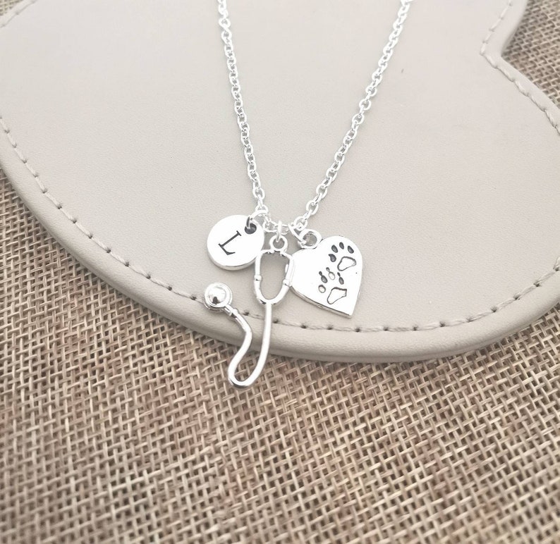 Peut inclure: Collier argent&eacute; avec un charme en forme de st&eacute;thoscope, un c&oelig;ur avec des empreintes de pattes et un charme rond avec la lettre "L". Le collier est pr&eacute;sent&eacute; sur une surface gris clair, sur un fond textur&eacute;.