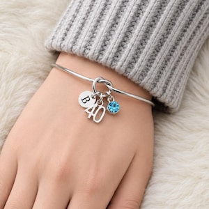 May include: A silver bangle bracelet with a knot design. The bracelet has three charms: a disc with the letter "B", the number "40", and a blue crystal. The bracelet is worn on a wrist.
