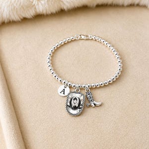 May include: Silver beaded bracelet with a cowboy hat, boot, and initial charm. The cowboy hat charm has a rope-like border. The bracelet is on a beige surface with a fluffy white background.
