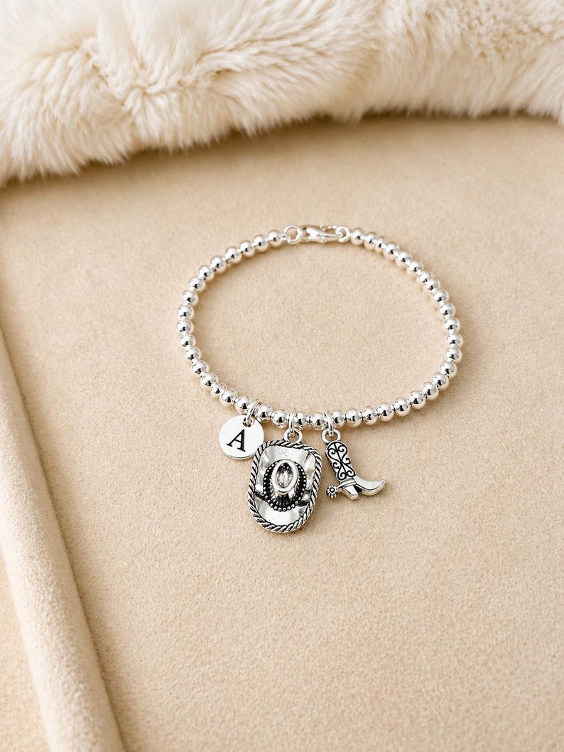 May include: Silver beaded bracelet with a cowboy hat, boot, and initial charm. The cowboy hat charm has a rope-like border. The bracelet is on a beige surface with a fluffy white background.