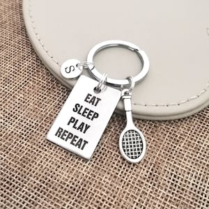 Op de afbeelding: Zilveren sleutelhanger met een tennisracket-bedel en een tag met de tekst "Eten Slapen Spelen Herhalen".