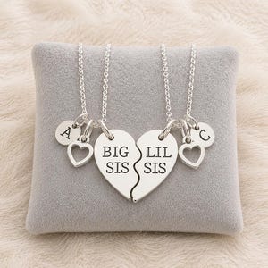 May include: Two silver necklaces with heart pendants, each with a chain. One pendant reads "BIG SIS" and the other "LIL SIS". Each necklace has a small heart charm and a round charm with a letter. The necklaces are displayed on a gray pillow.
