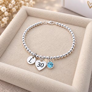 Op de afbeelding: Een zilverkleurige kralenarmband met drie bungelende bedels: een ronde schijf gegraveerd met de letter L, een hartvormige bedel gegraveerd met het getal 30 en een kleine ronde bedel met een enkele helderblauwe edelsteen.