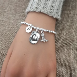 May include: Silver beaded bracelet with a cowboy hat charm, a cowboy boot charm, and a round charm with the letter "R". The bracelet is on a wrist, with a gray sweater sleeve in the background.