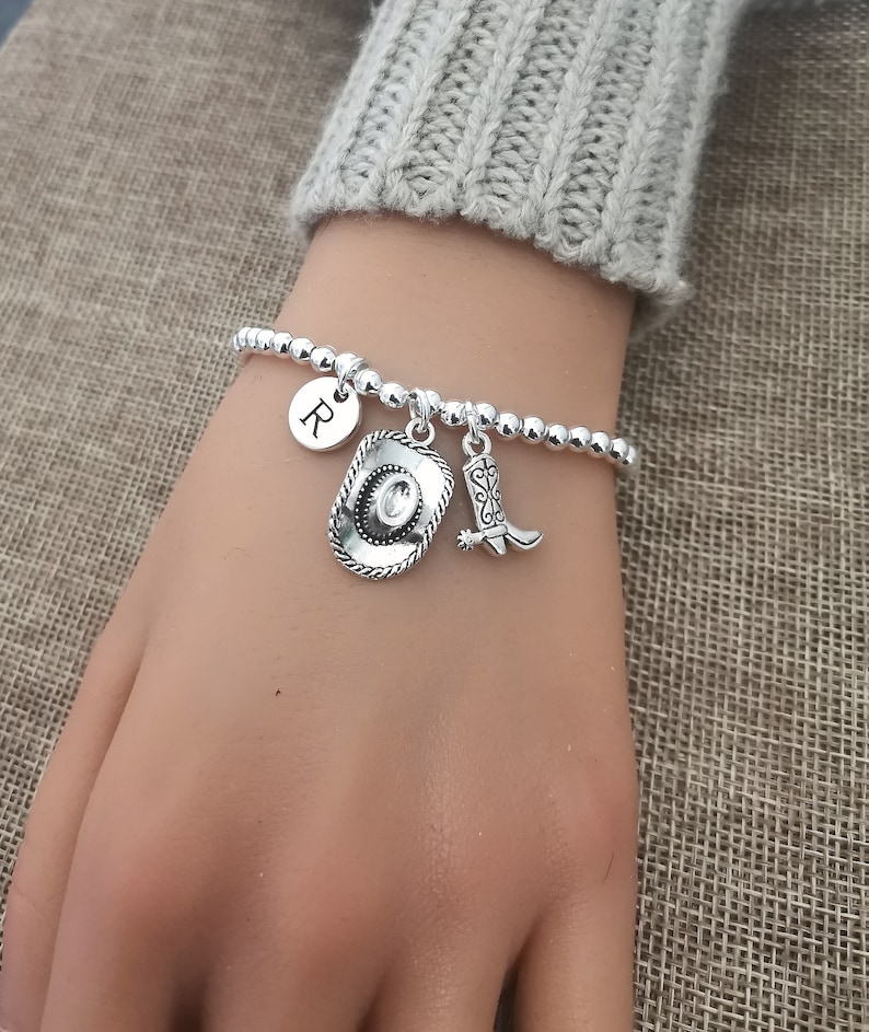 May include: Silver beaded bracelet with a cowboy hat charm, a cowboy boot charm, and a round charm with the letter "R". The bracelet is on a wrist, with a gray sweater sleeve in the background.