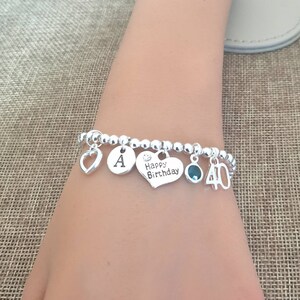 May include: Silver charm bracelet with a heart, letter A, heart with the text "Happy Birthday", a green gemstone, and a charm with the number 40.
