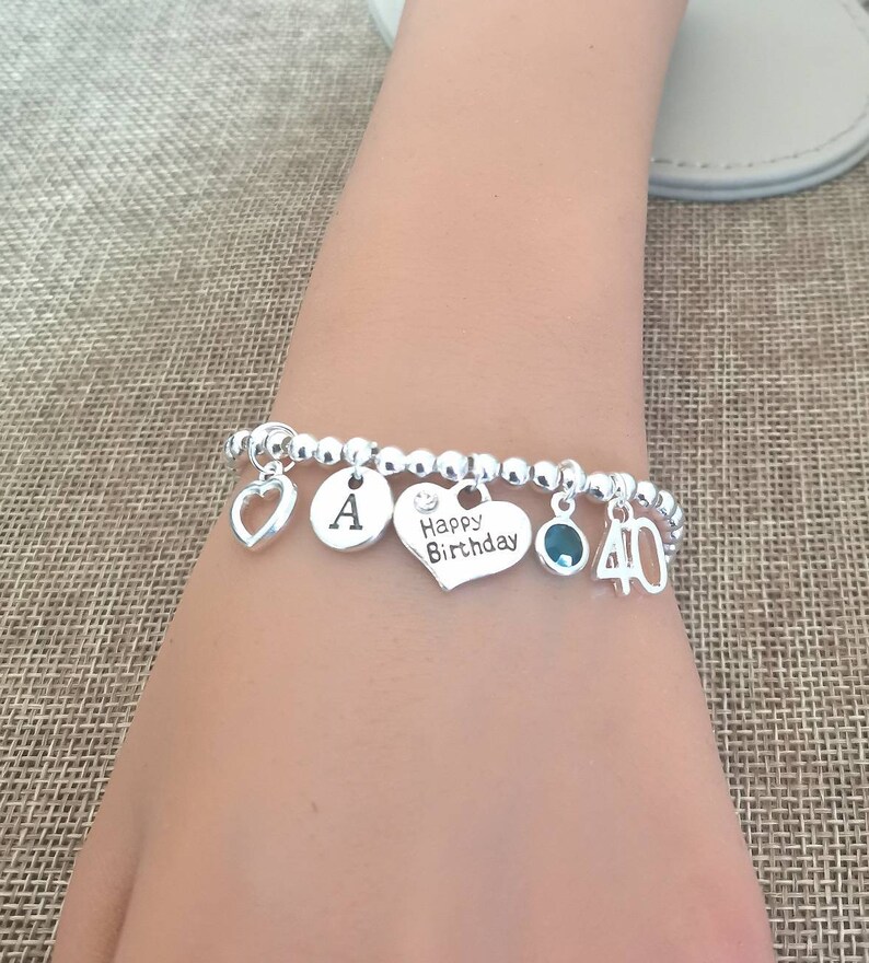 May include: Silver charm bracelet with a heart, letter A, heart with the text "Happy Birthday", a green gemstone, and a charm with the number 40.