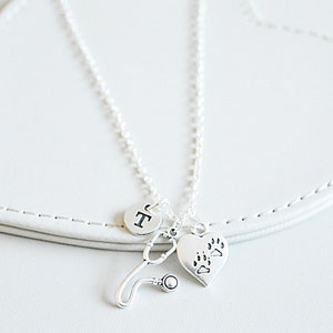 Peut inclure: Un collier argent&eacute; avec un charme de st&eacute;thoscope, un charme en forme de c&oelig;ur avec des empreintes de pattes et un charme rond avec la lettre "T". Le collier est pr&eacute;sent&eacute; sur une surface blanche en forme de c&oelig;ur.